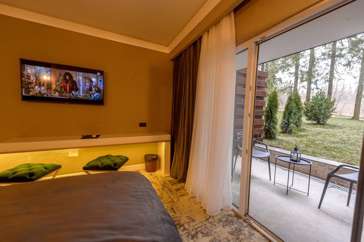 A hotel room with a bed, a television, a balcony with chairs and a table, and a view of trees and grass.