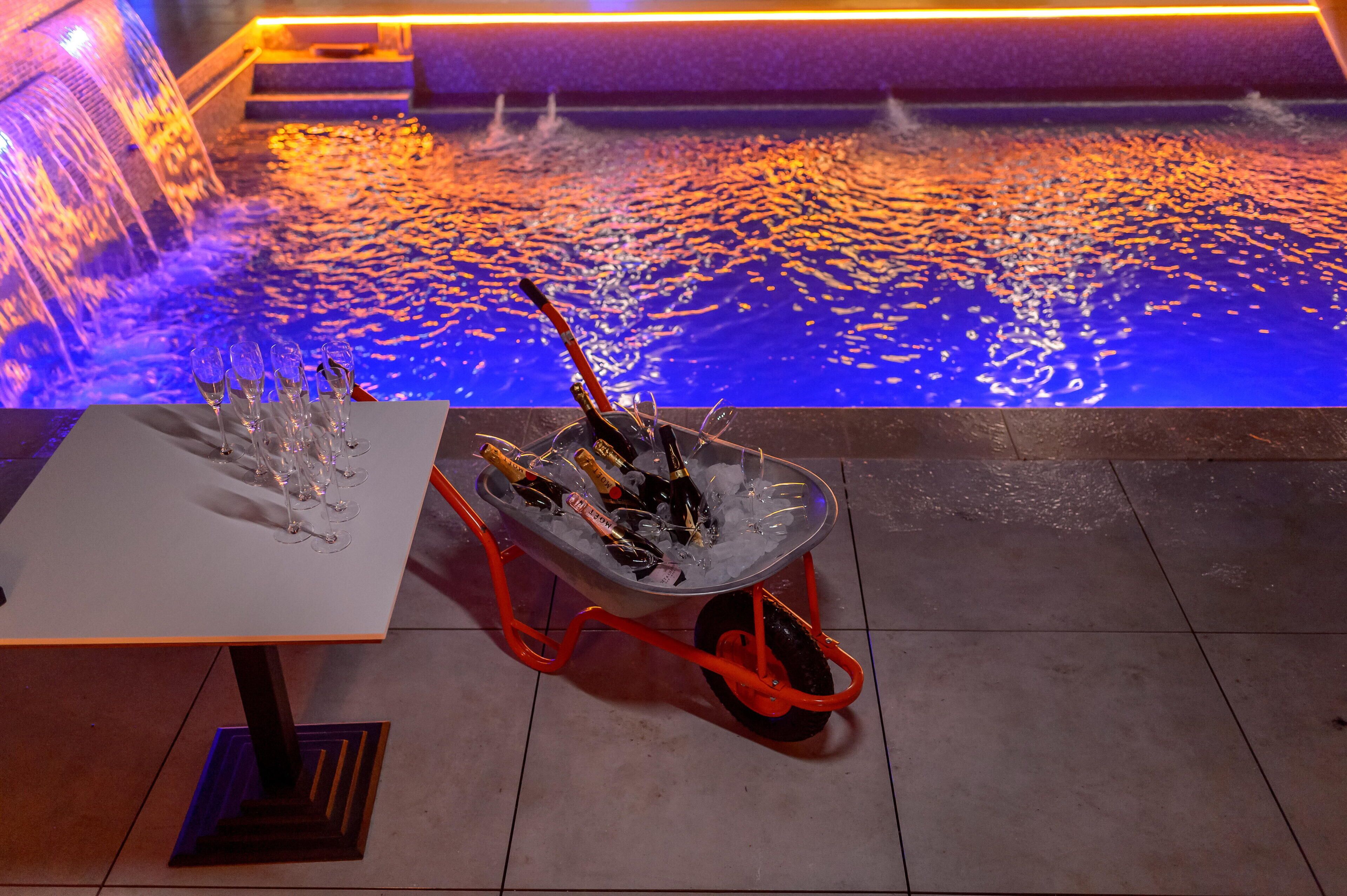 A pool with a water feature and a table with a cart of bottles and glasses.
