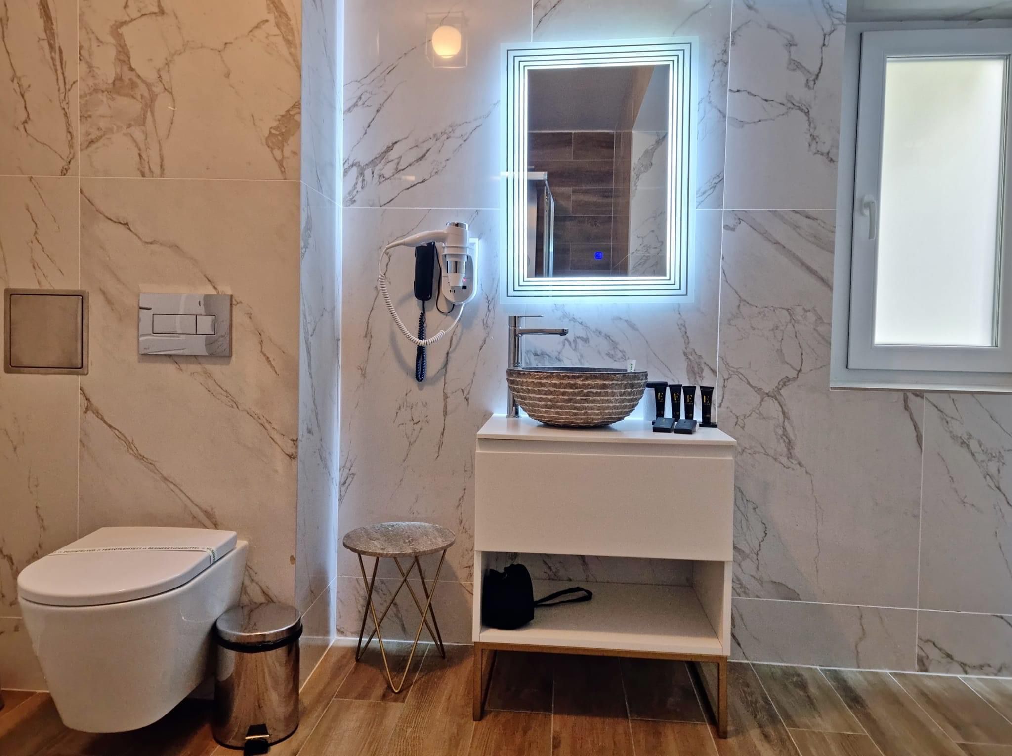 A modern bathroom with a marble wall, a white sink with a vessel basin, a small stool, and a toilet.
