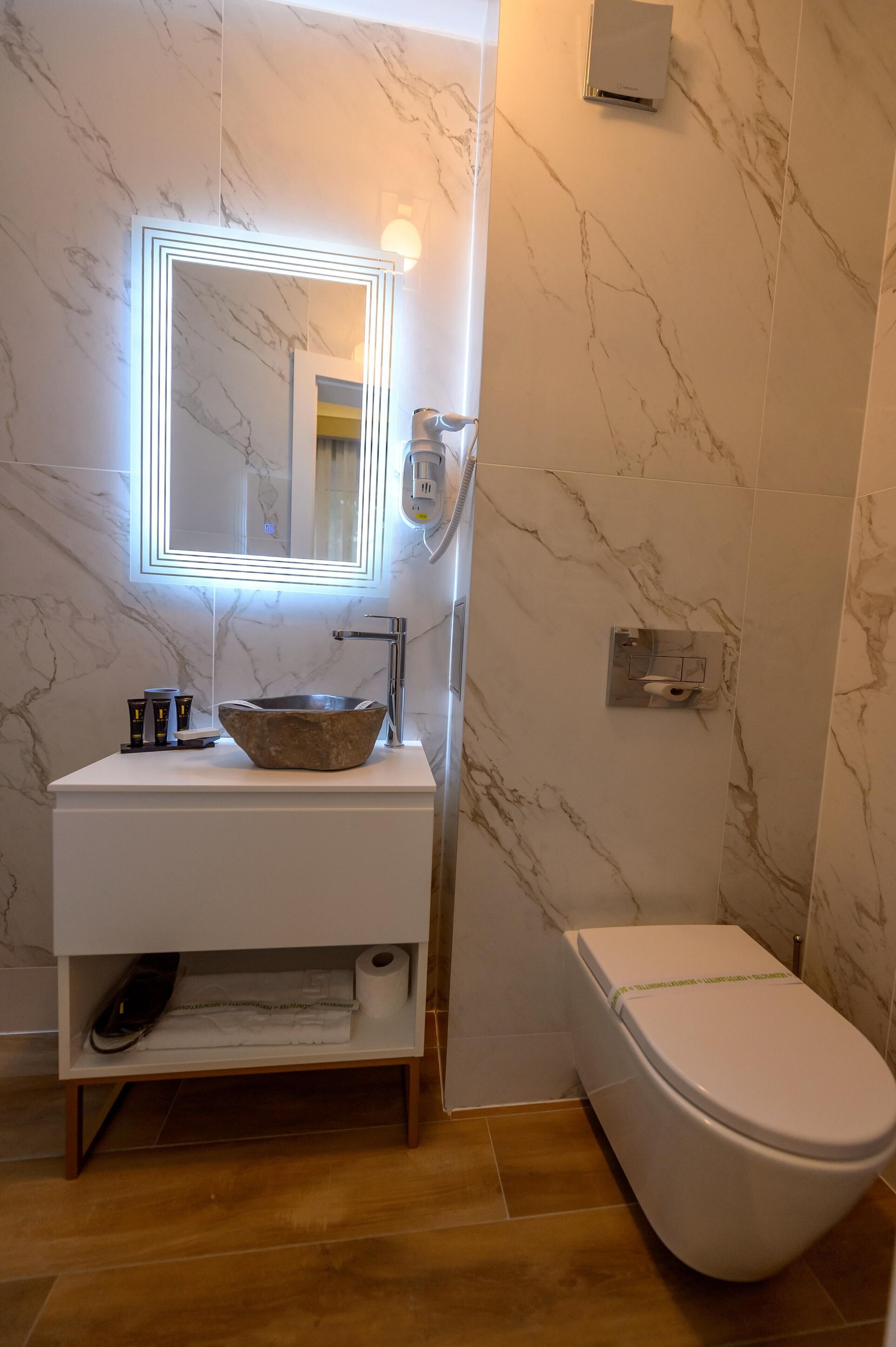 Un baño con pared de mármol, lavabo blanco, espejo grande y un inodoro.