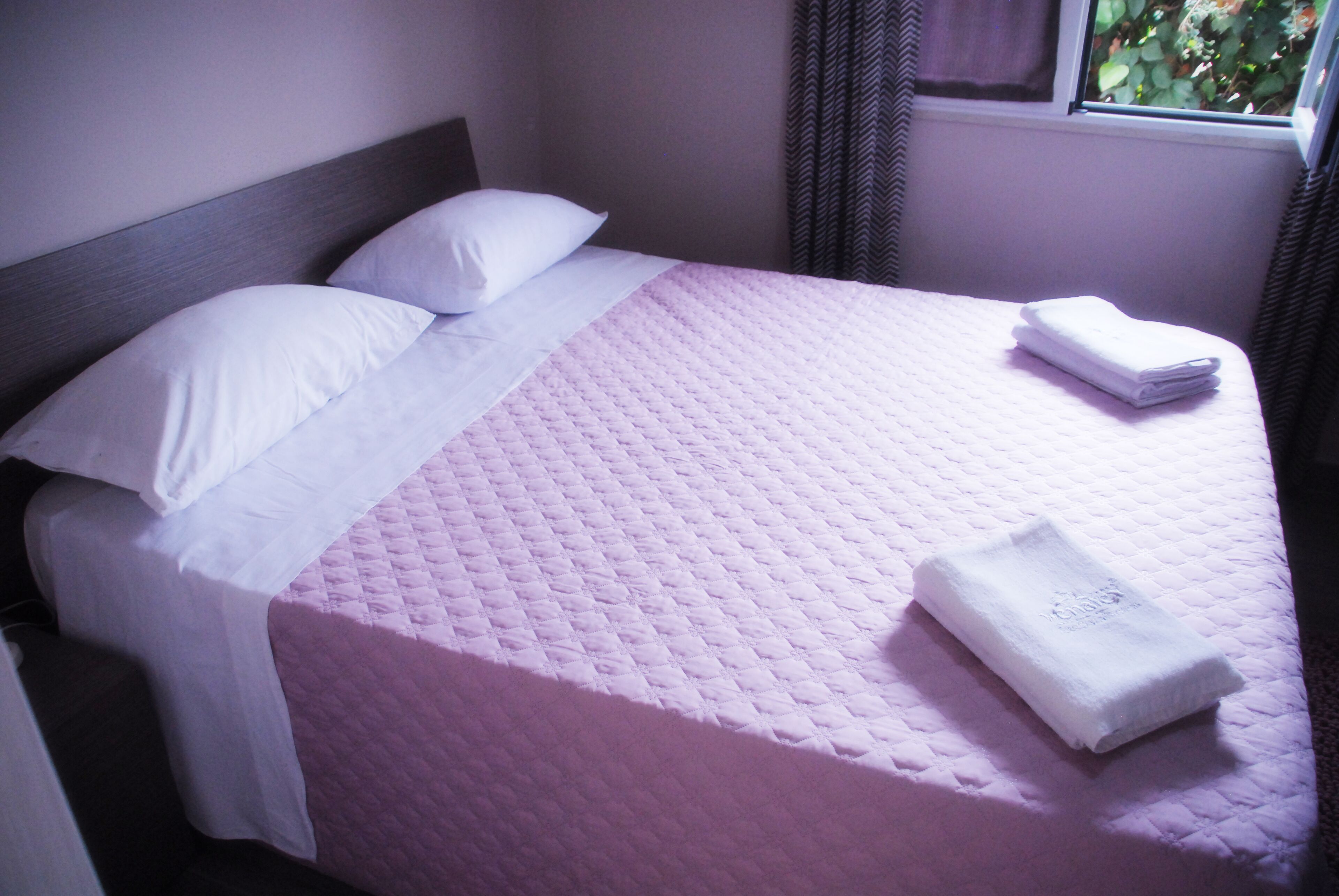 Ein ordentlich bezogenes Bett mit einer rosa Quilttdecke, weißen Kissen und einem gefalteten weißen Handtuch an der Seite.