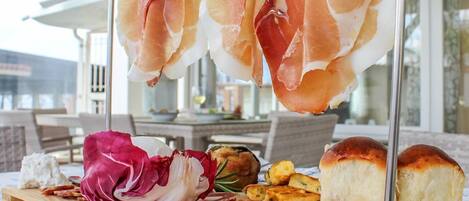 Una tabla de madera con diversos alimentos, como jamón serrano, queso y pan, expuesta al aire libre.