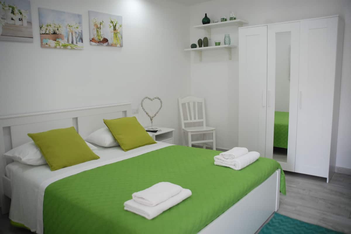 Une chambre à coucher bien rangée, avec un lit, une literie verte et blanche, une décoration en forme de cœur, une chaise blanche et une armoire.
