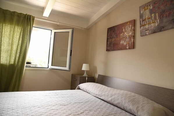 A bedroom with a bed, a nightstand with a lamp, a window with curtains, and two wall art pieces. - Marechiaro Paestum (Capaccio-Paestum)