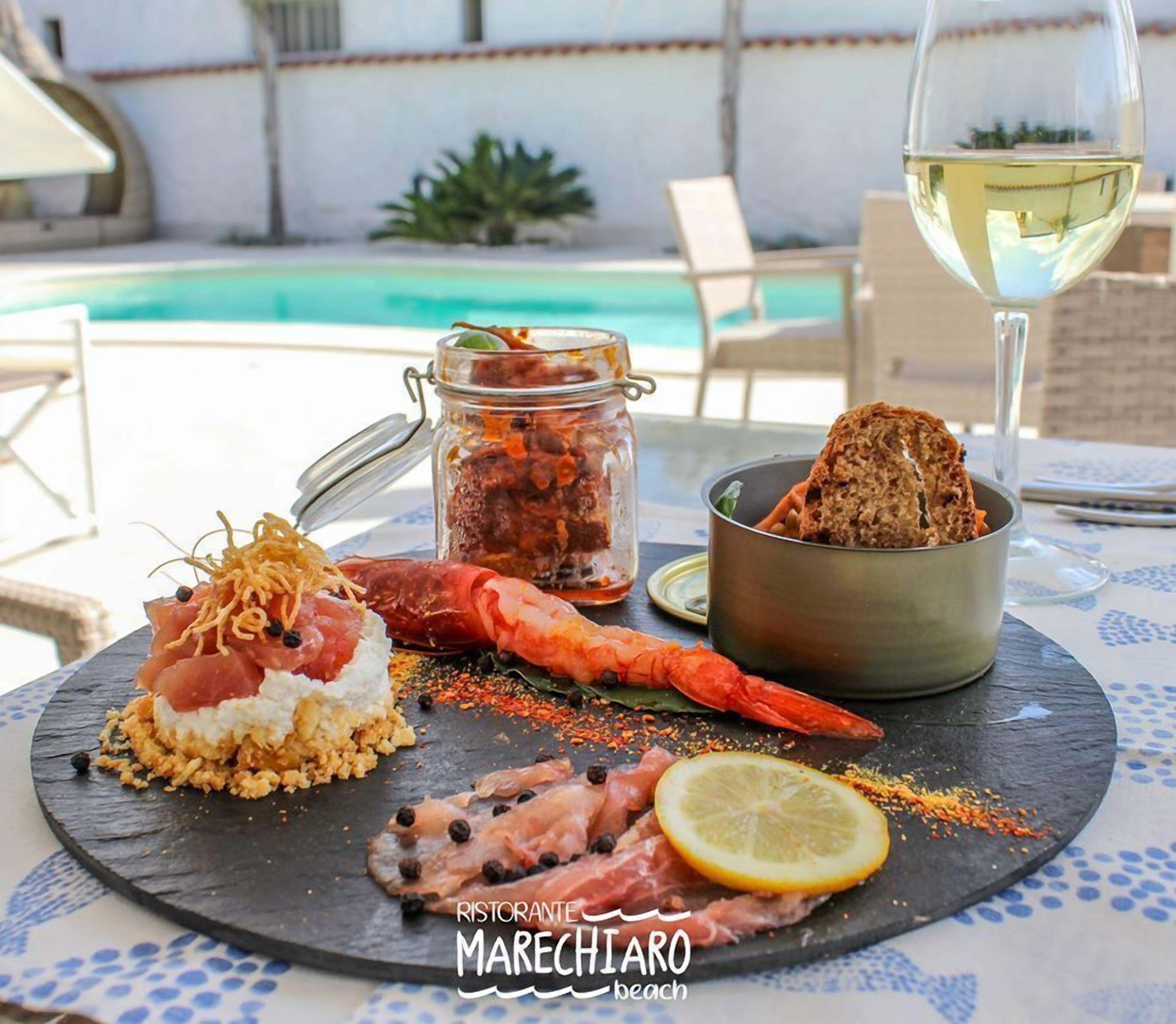 A seafood platter with shrimp, fish, and bread on a slate board, accompanied by a glass of white wine and a jar of sauce, set on a table with a pool view in the background.