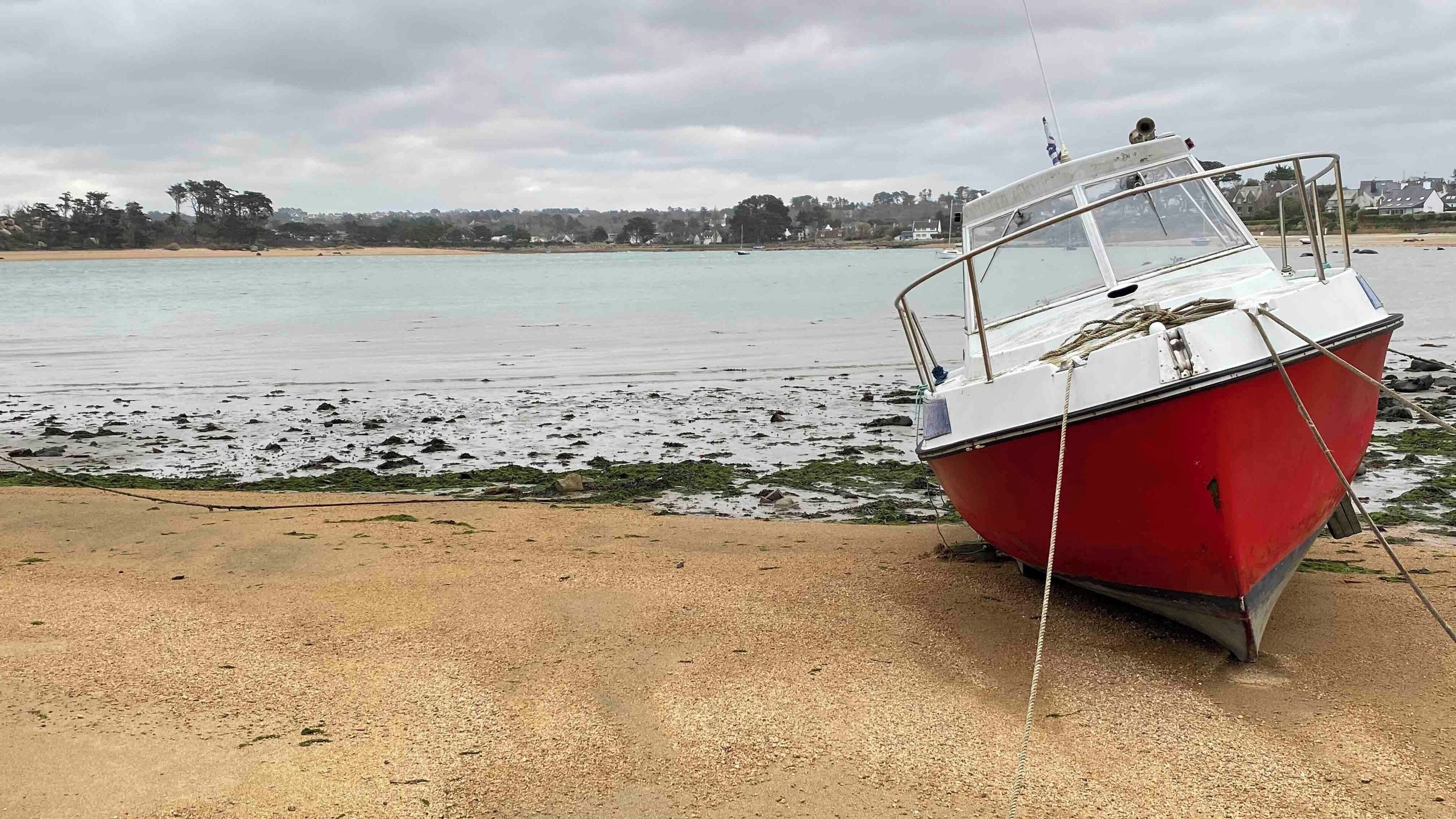 A red boat tied to a sandy beach with calm water and a cloudy sky in the background.