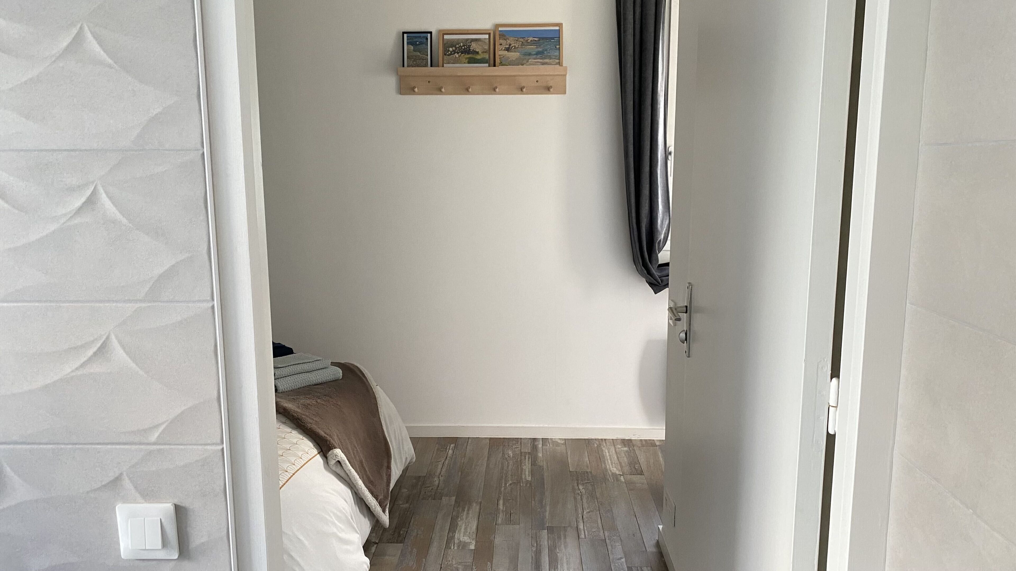 Kapea käytävä, jonka päässä on sänky, seinälle kiinnitetty naulakko, jossa on takki, ja seinälle asennettu hylly, jossa on valokuvia.