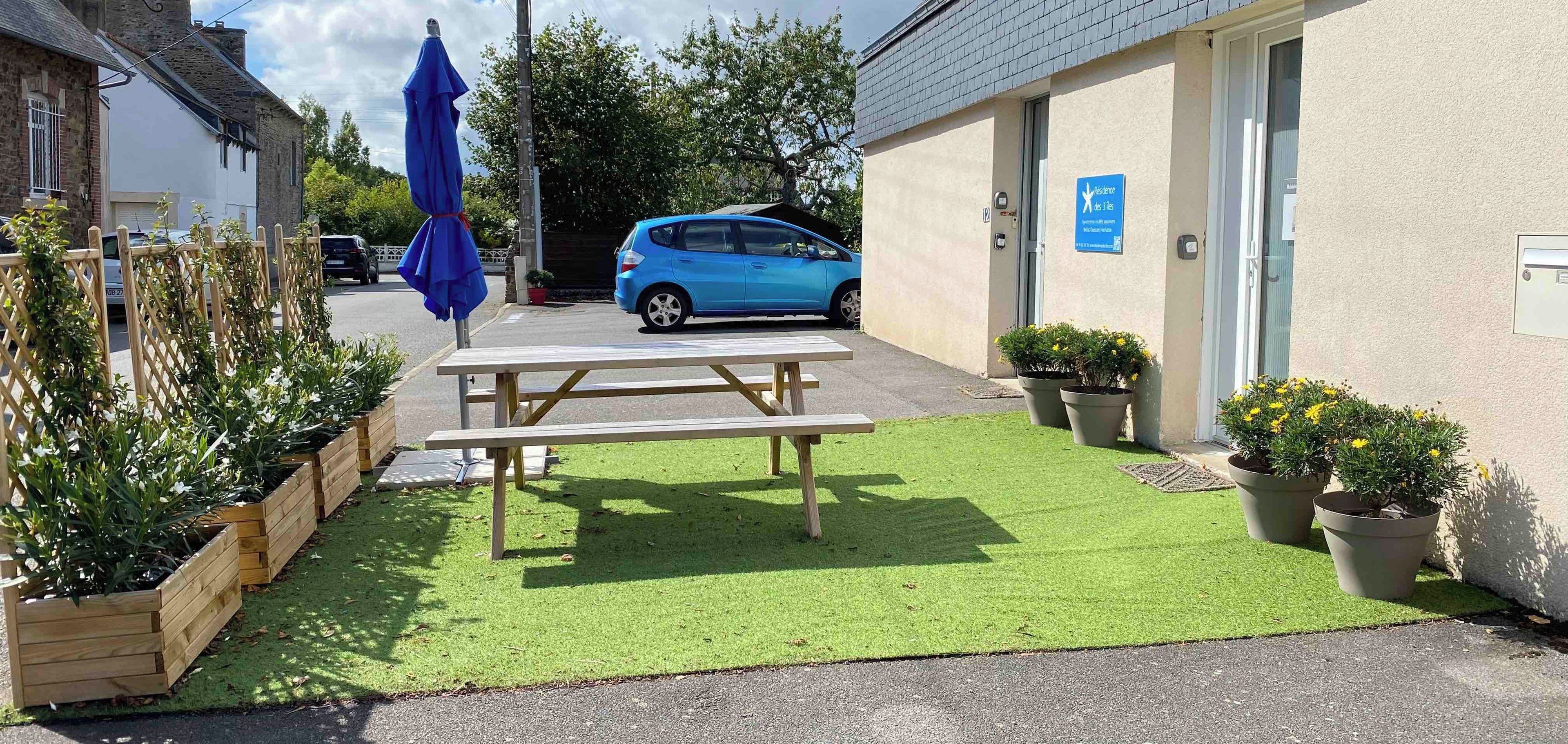 Une cour avec une table de pique-nique, une voiture bleue garée à proximité et un bâtiment portant une enseigne indiquant qu’il s’agit d’un hôtel trois étoiles.