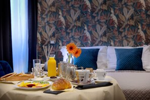 Una mesa de desayuno con diversos alimentos, un florero con una flor y una cama con almohadas al fondo.