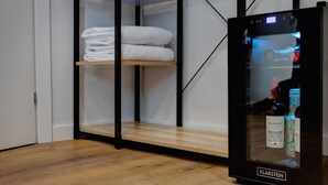 Una habitación moderna con suelo de madera, un mini bar con puerta de vidrio y un estante ordenado con toallas dobladas.