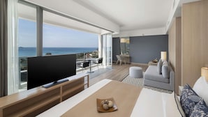 A modern hotel room with a large TV, a sofa, a dining table, and a balcony with a view of the sea.