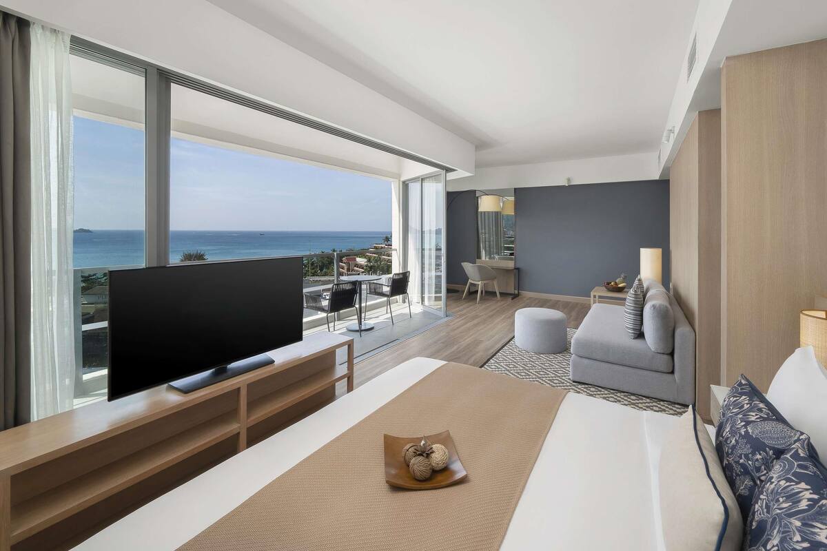 A modern hotel room with a large TV, a sofa, a dining table, and a balcony with a view of the sea.