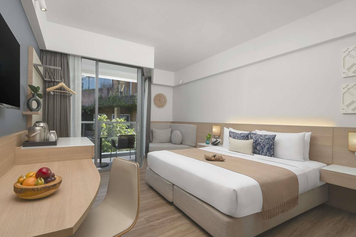 A modern hotel room with a bed, bedside table, and a dining area with a fruit bowl.