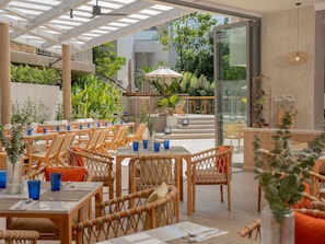 An outdoor dining area with wicker furniture and blue glassware, surrounded by greenery and a glass door leading to a balcony. - Andaman Beach Hotel Phuket - Handwritten Collection by Accor (Patong)
