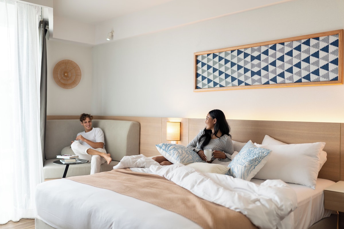 A hotel room with a bed, a sofa, a wall-mounted artwork, and a bedside table with a lamp.