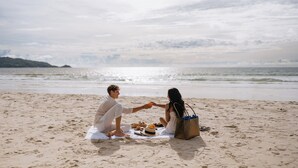 A couple sitting on a beach towel with a picnic setup, including food and drinks, and a beach bag. - Andaman Beach Hotel Phuket - Handwritten Collection by Accor (Patong)