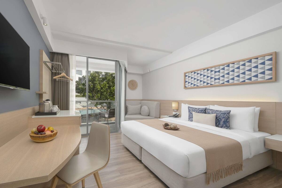 A modern hotel room with a large bed, a desk, and a balcony with a pool view.