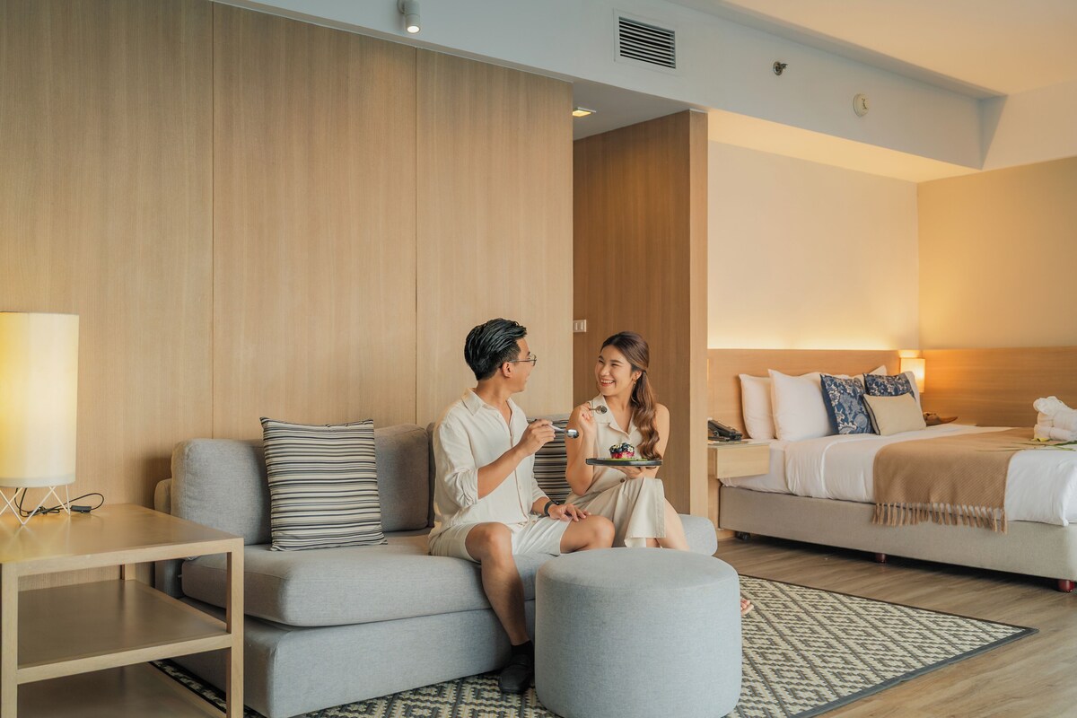 A modern hotel room with a sofa, ottoman, bedside table, and a bed with pillows and a blanket.