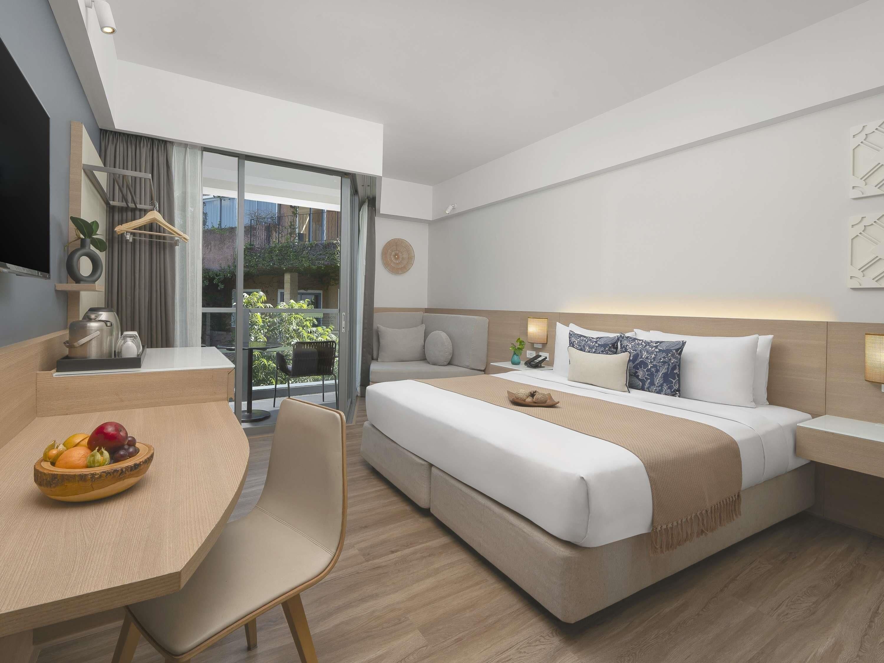 A modern hotel room with a bed, bedside table, and a dining area with a fruit bowl.