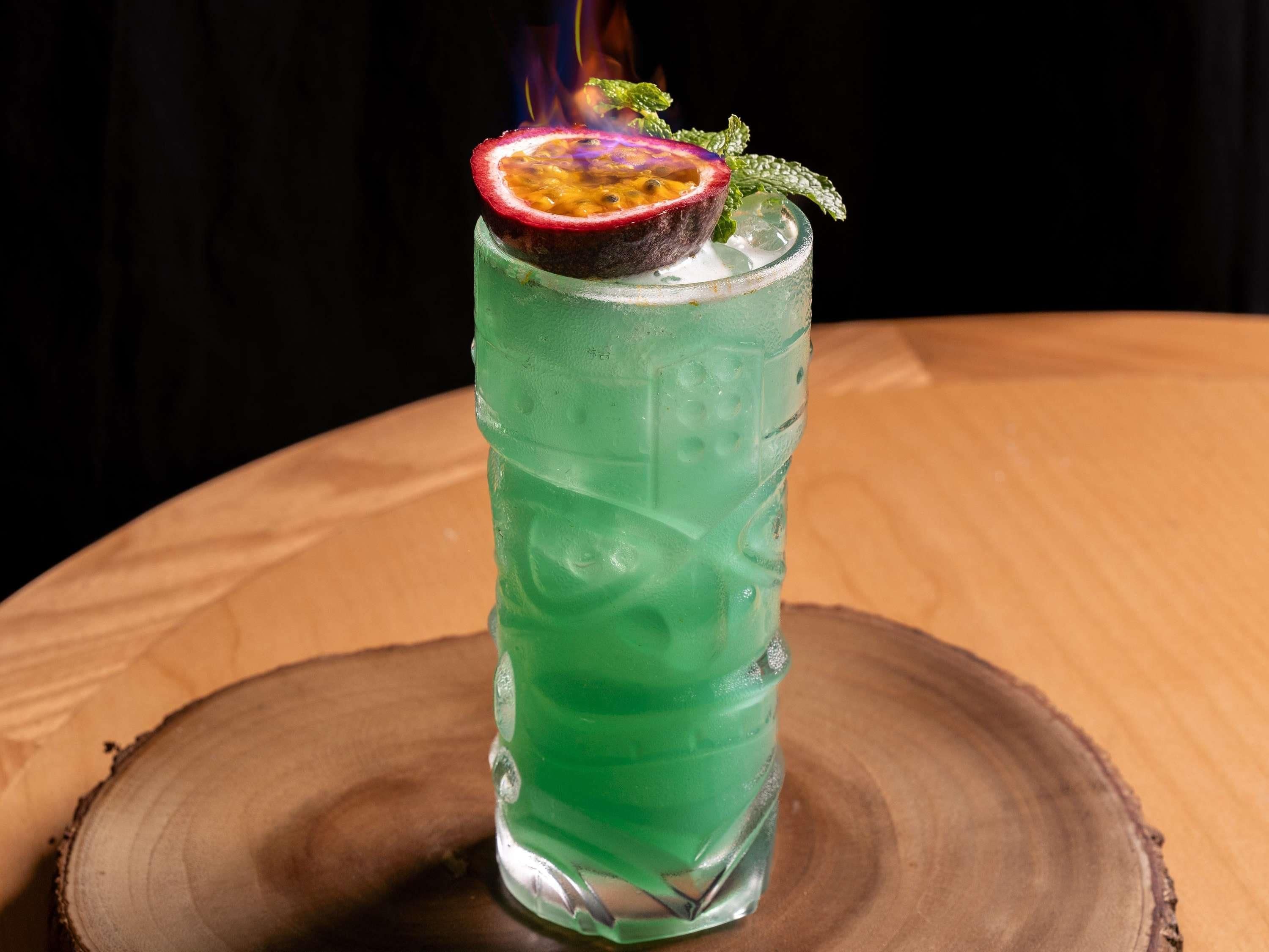 Un vaso alto con un líquido verde, una tajada de maracuyá y una ramita de menta sobre una superficie de madera.