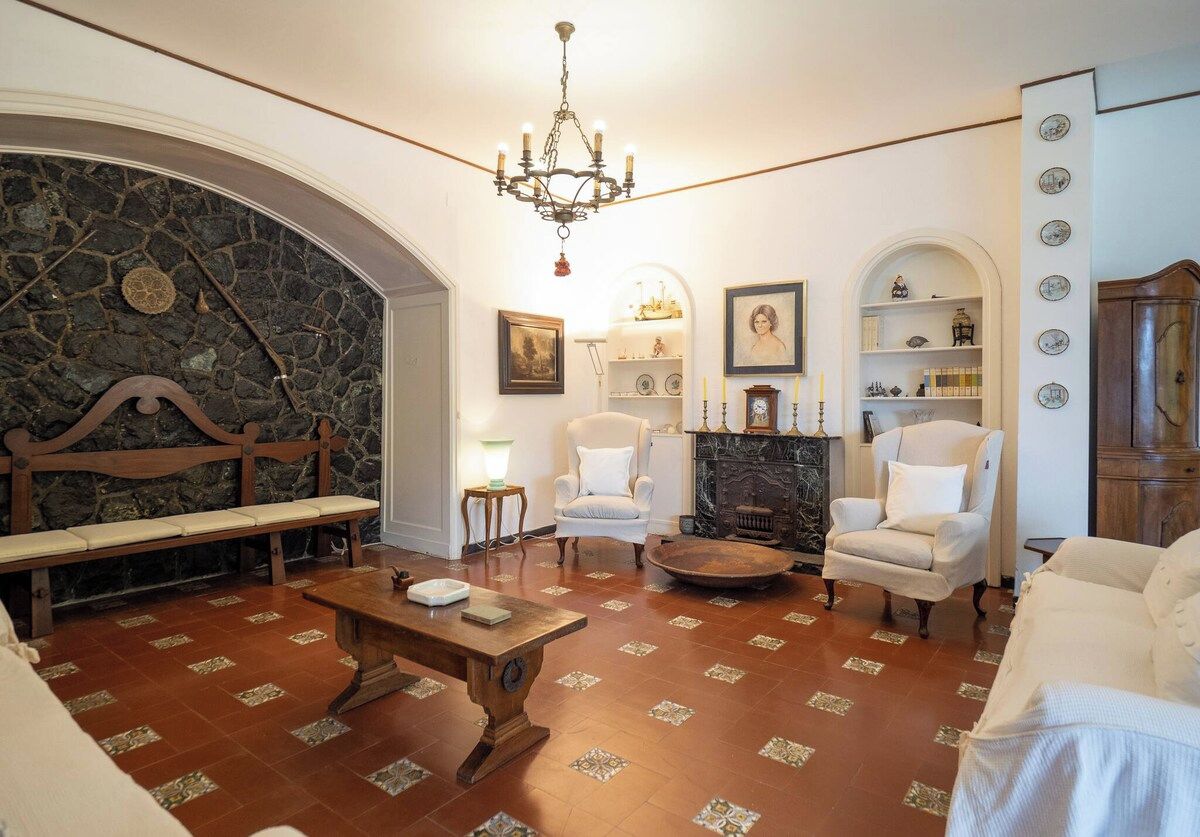 A traditional living room with a stone fireplace, white armchairs, a wooden bench, and a chandelier.