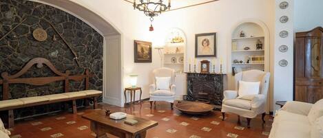 Un salon traditionnel avec une cheminée en pierre, des fauteuils blancs, un banc en bois et un lustre.
