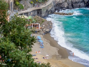 A coastal beach with a rocky cliff, a wooden walkway, and a small building with a beach umbrella.