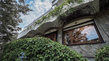 A building with stone walls and large windows, surrounded by lush greenery.