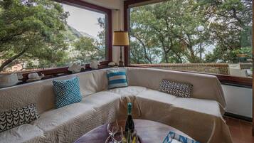 A room with a large window, a sofa, a table with a bottle and glasses, and a view of trees and mountains.