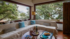 A room with a large window, a sofa with patterned cushions, a dining table with a tray of food, and a bottle of wine. - Villa Tanca with Terrace and Views (Monterosso al Mare)