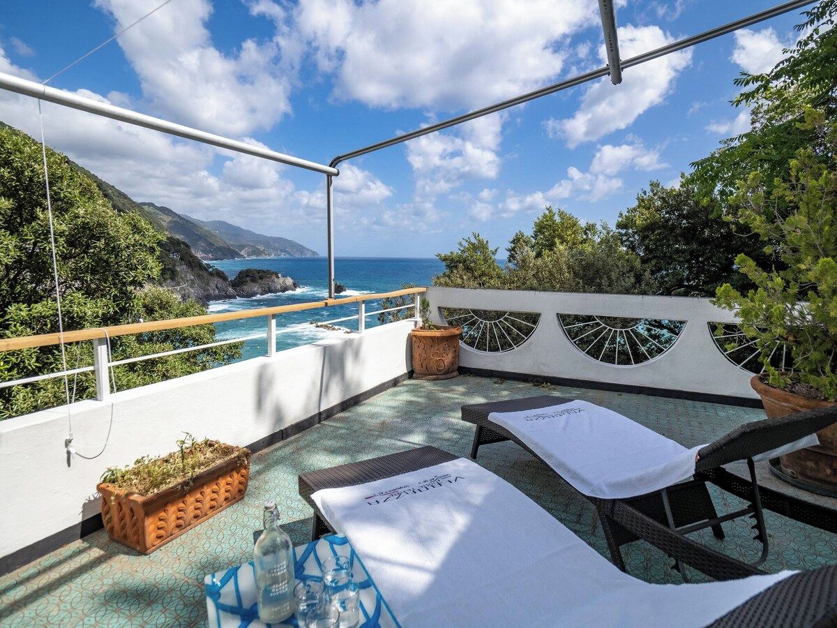 A rooftop terrace with a view of the sea, lounge chairs, and a small table with a bottle.