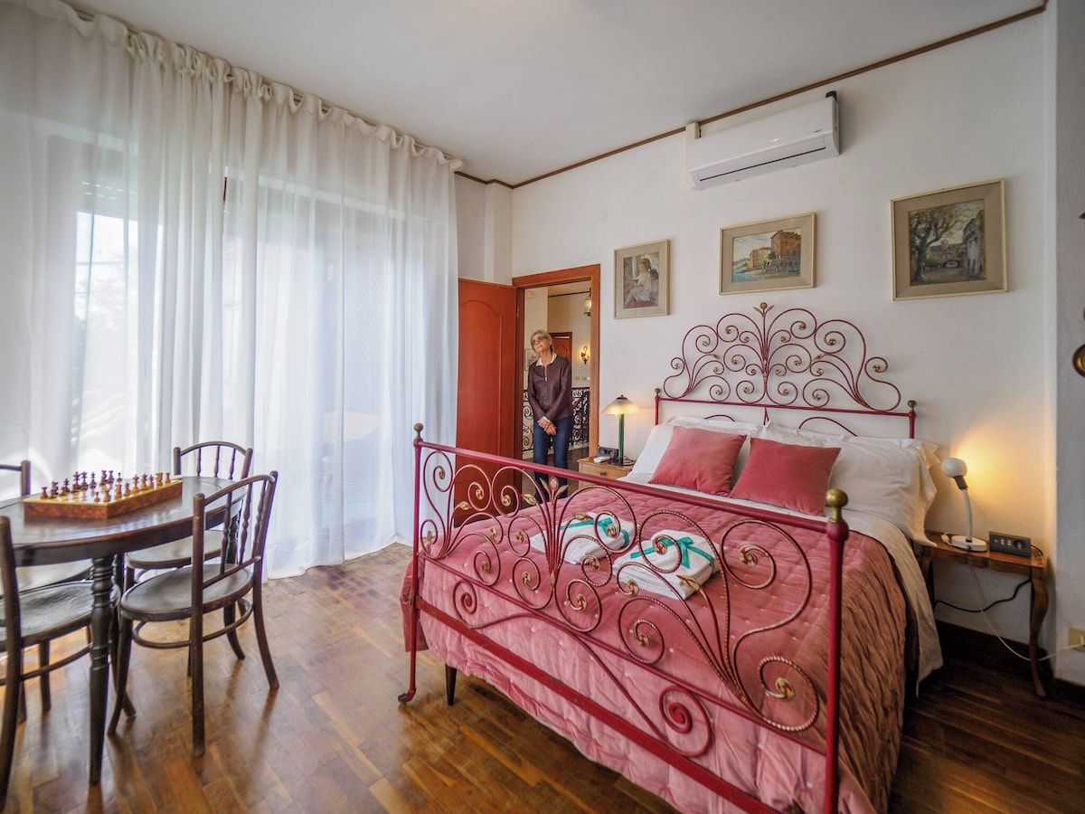 A bedroom with a bed, a bedside table, a lamp, a chair, a table with a chessboard, and a window with curtains.
