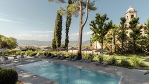 A swimming pool with lounge chairs and umbrellas, surrounded by greenery and a view of a historic building. - Grand Hotel San Gemini | UNA Esperienze. (San Gemini)