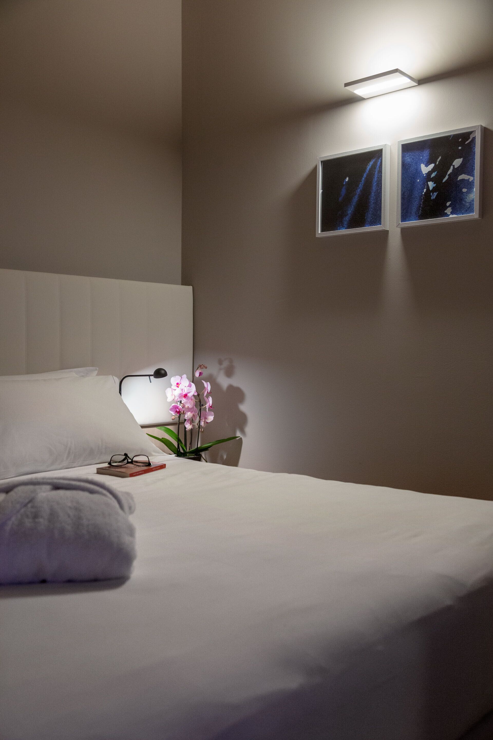 A hotel room with a neatly made bed, a bedside lamp, a small table with a vase of flowers, and two framed pictures on the wall.