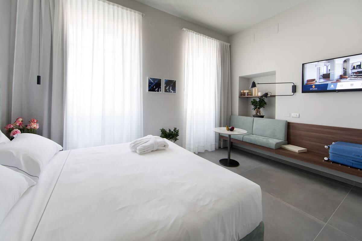 Una habitación de hotel con una cama grande, una mesita con una planta, un sofá y un televisor fijado a la pared.
