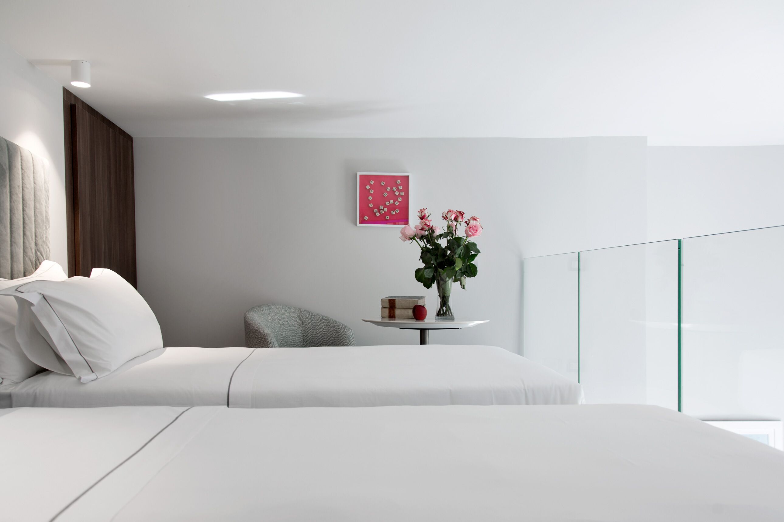 A modern hotel room with a large bed, a small round table with a vase of flowers, a framed picture on the wall, and a glass railing.