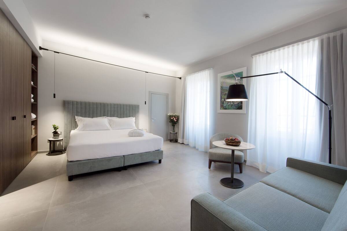 Una habitación de hotel moderna con una cama grande, un sofá, una mesita con un jarrón y una lámpara.