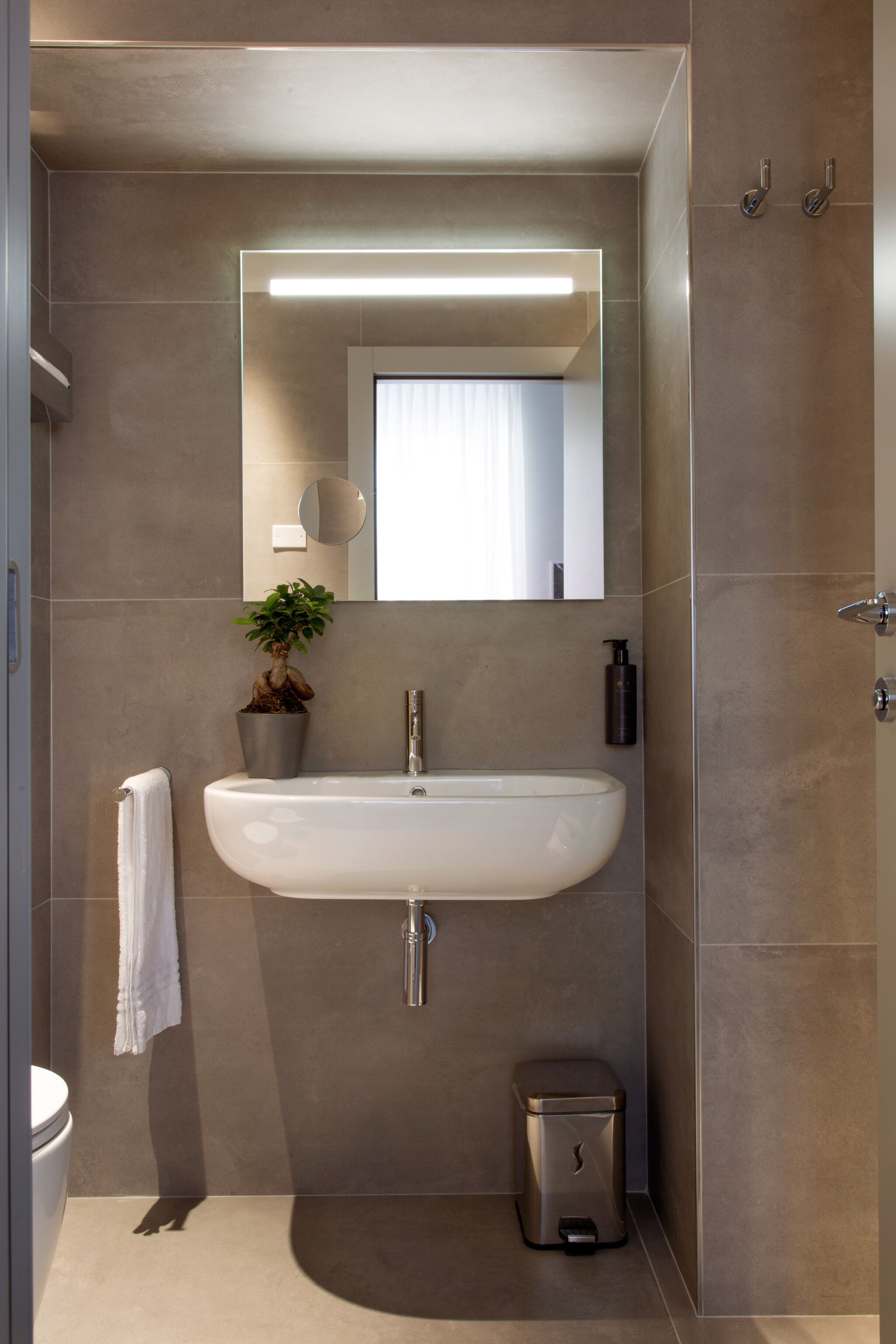 Un baño moderno con lavamanos blanco, un espejo, una planta en maceta, una toalla y un cubo de basura.