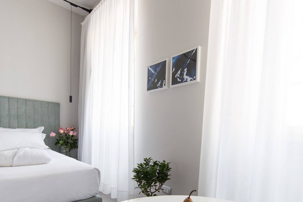 Una habitación de hotel moderna con una cama bien hecha, una mesita de mármol con frutas, una planta pequeña en maceta y una obra de arte en la pared.