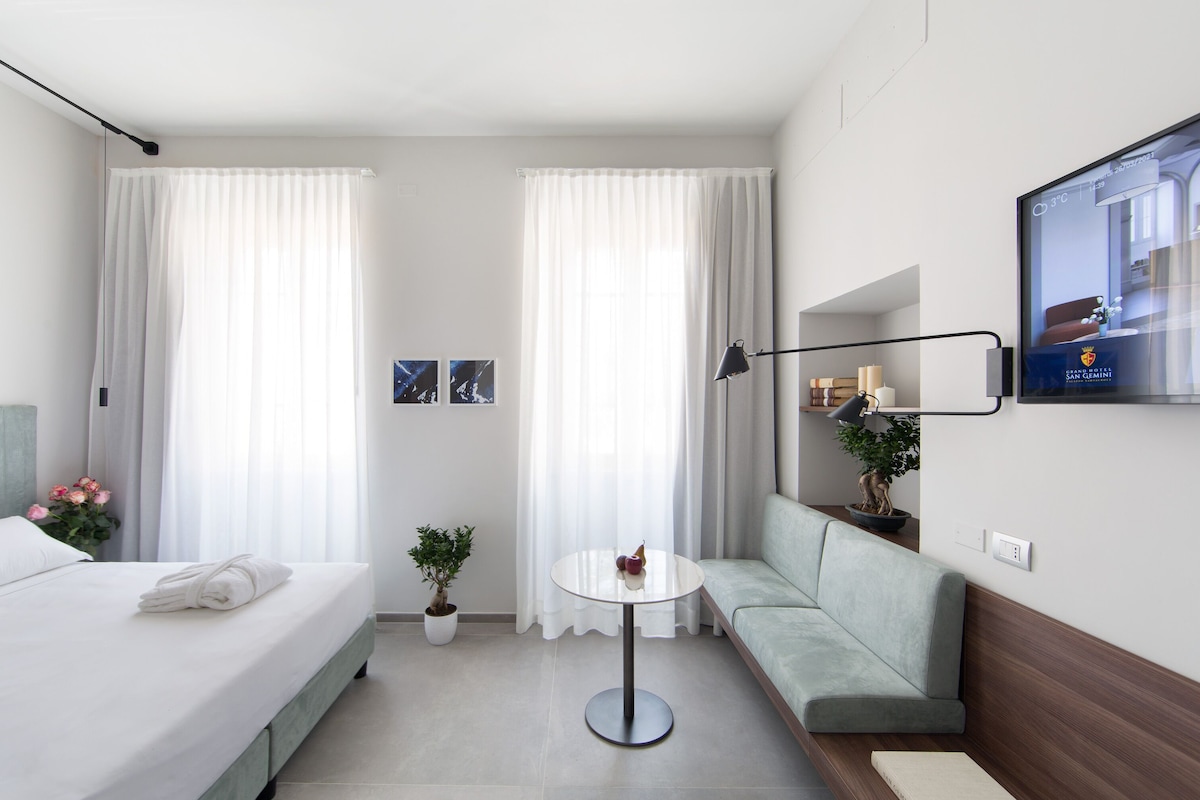 A modern hotel room with a bed, a sofa, a small table, a potted plant, and a TV on the wall.