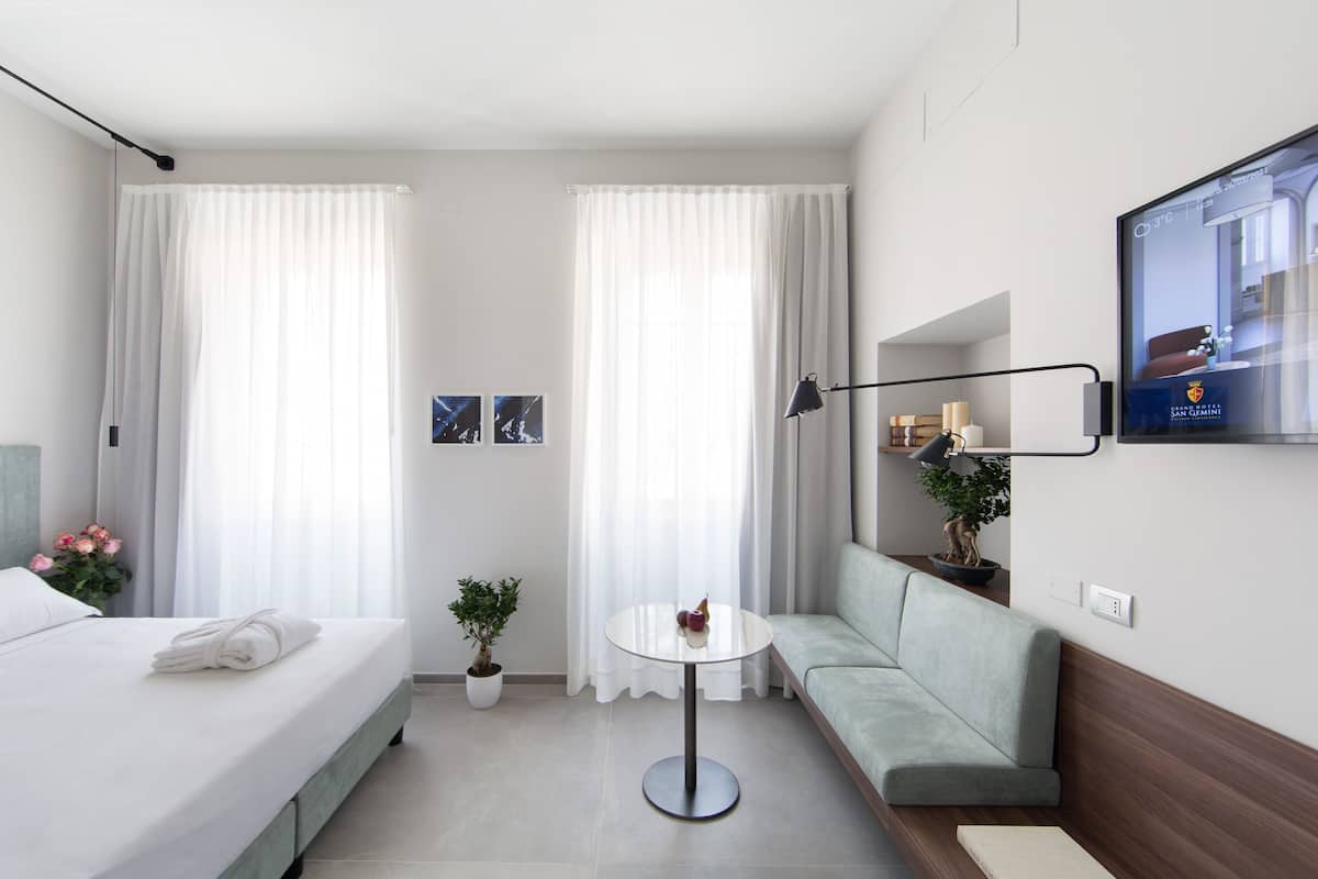A modern hotel room with a bed, a sofa, a small table, a potted plant, and a TV on the wall.