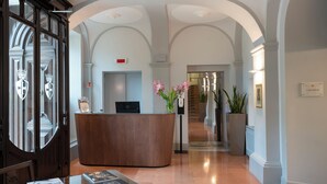 A reception area with a wooden reception desk, a computer, and a vase with pink flowers. - Grand Hotel San Gemini | UNA Esperienze. (San Gemini)