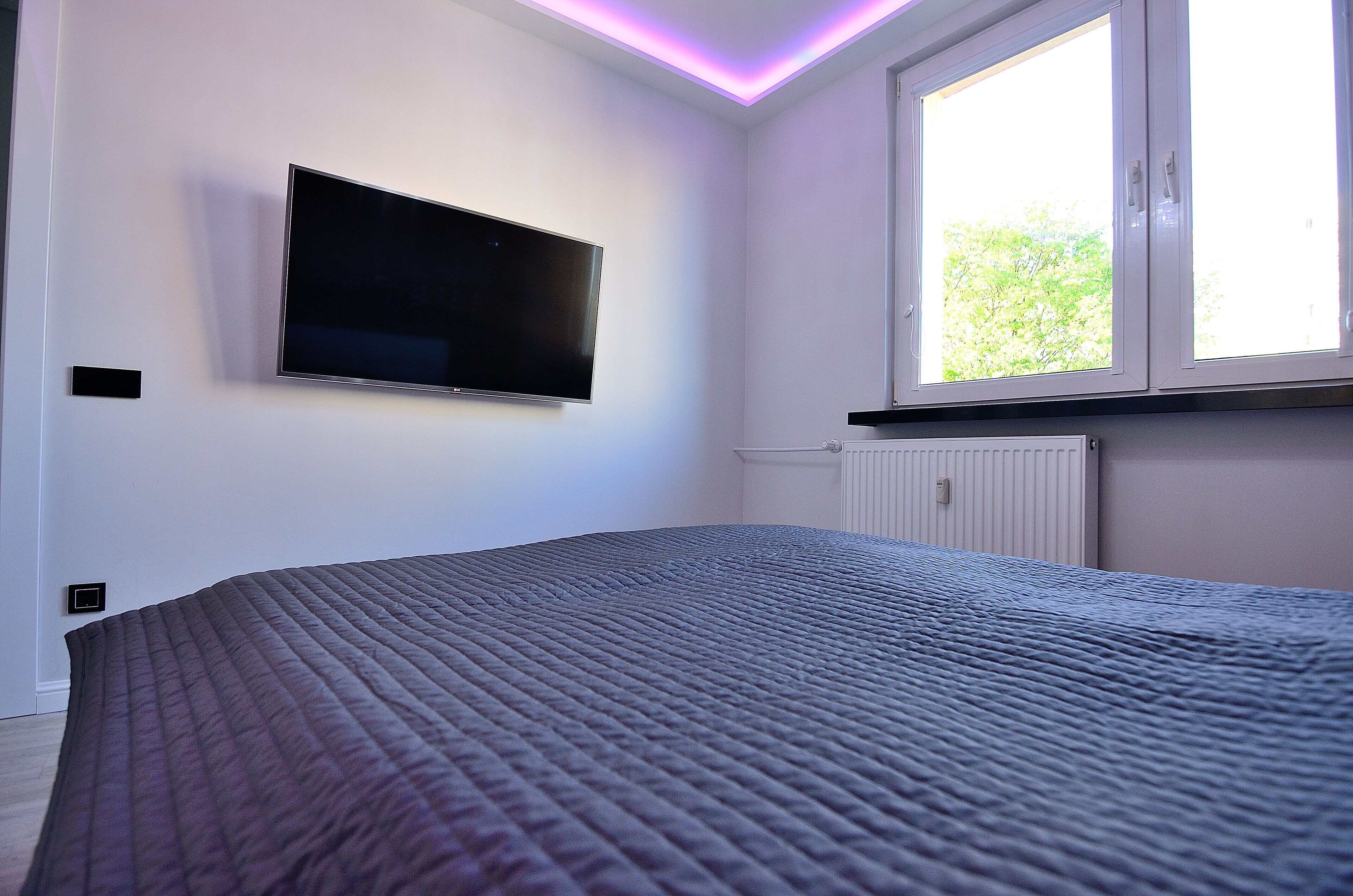A bedroom with a bed, a mounted TV, a window with blinds, and a radiator.