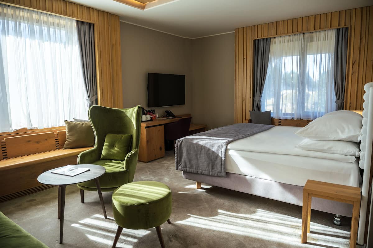 A hotel room with a bed, a green armchair, a small table, a desk with a TV, and large windows with curtains.