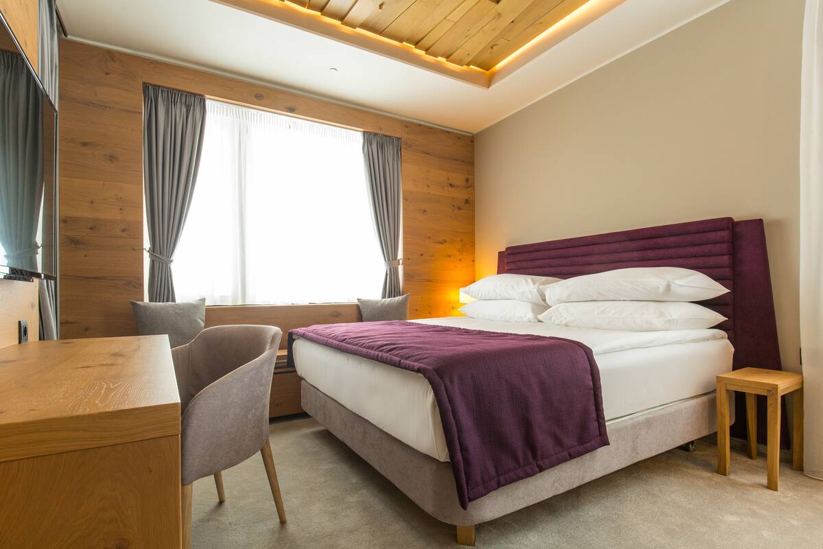 A bedroom with a wooden ceiling, a bed with white linens and a purple headboard, a wooden desk, and a chair.