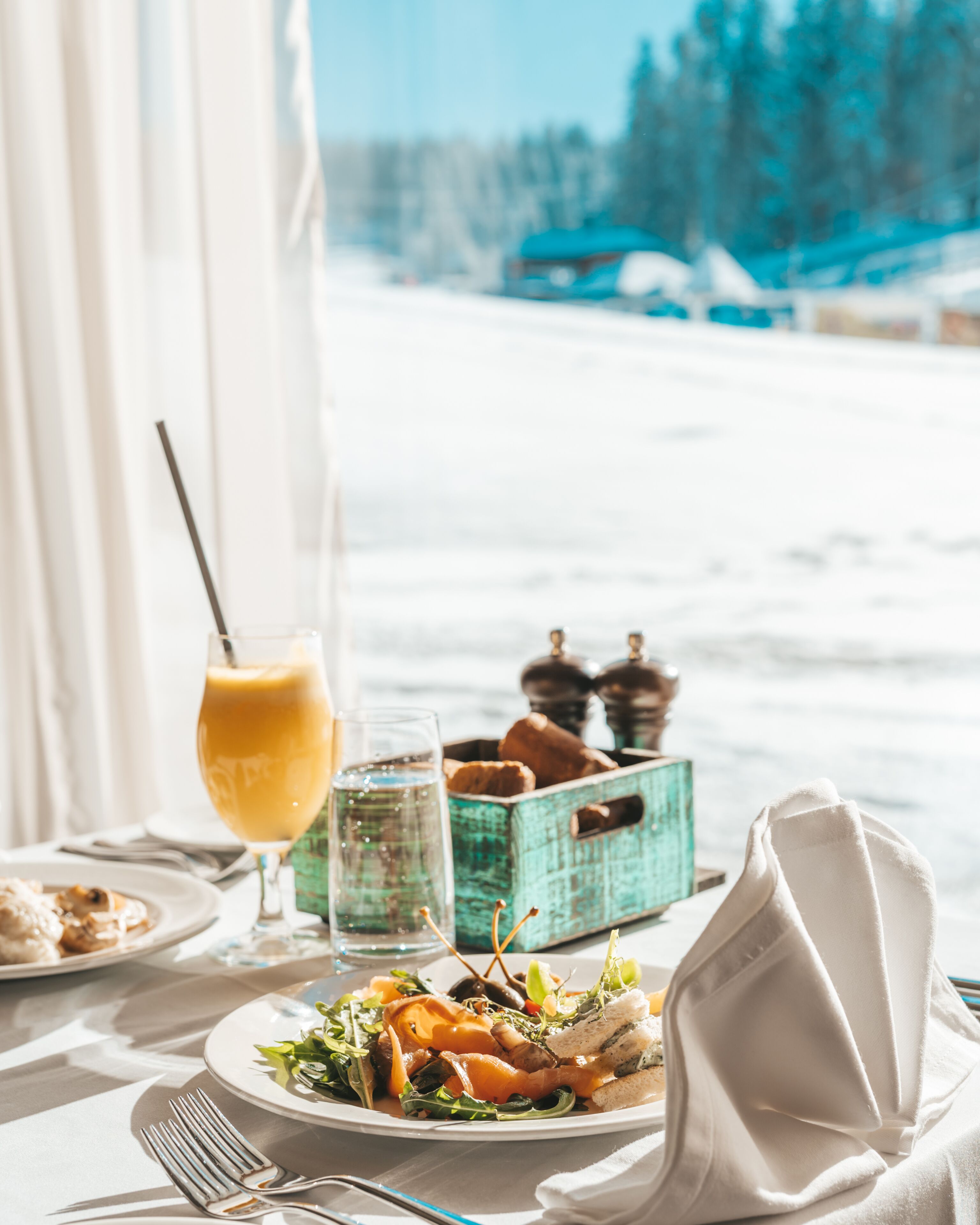 Una mesa de comedor con comida, un vaso de jugo de naranja y un vaso de agua. Se ve una ventana con vistas a un paisaje nevado y un edificio al fondo.