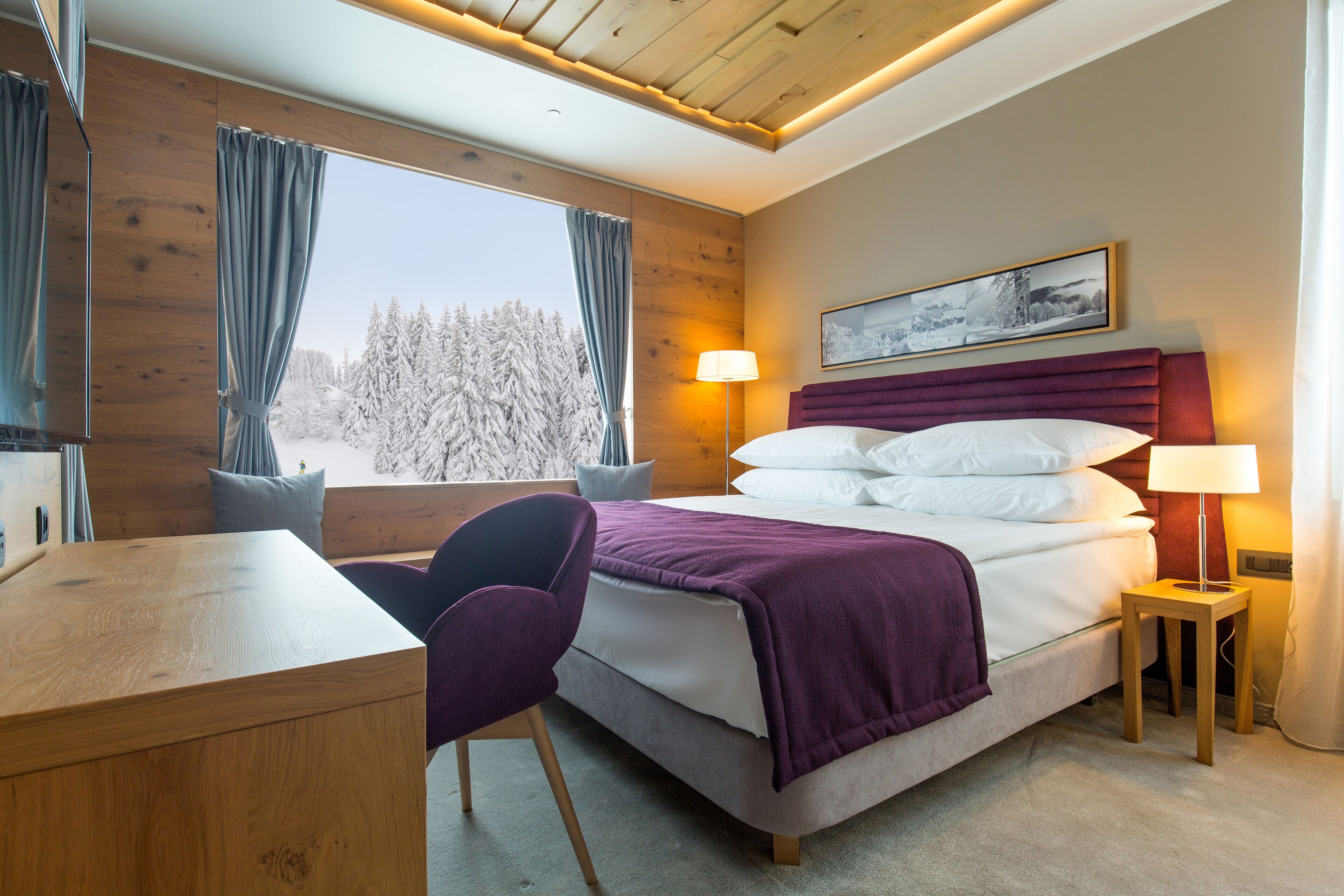 A modern hotel room with a wooden ceiling, a large bed with white linens, a purple blanket, a desk with a chair, and a window with a view of snow-covered trees.