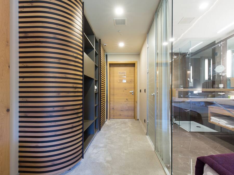Un pasillo moderno de hotel con un revestimiento de madera curva, un separador de vidrio y un área de baño visible con lavamanos y espejo.