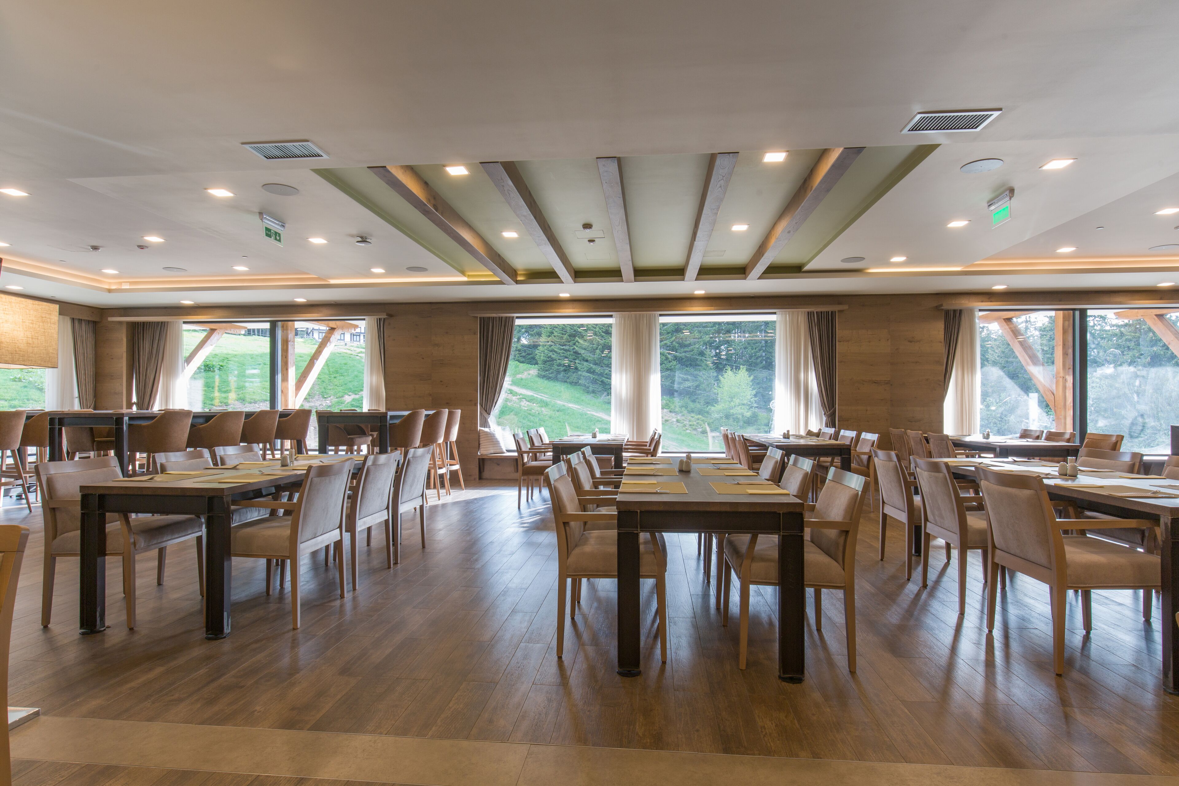 Un comedor espacioso con varias mesas y sillas, ventanas amplias con cortinas, y piso de madera.