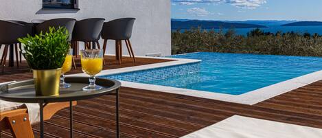 Zona junto a la piscina con terraza de madera, una mesa con planta y bebidas, y vistas al mar y montañas.
